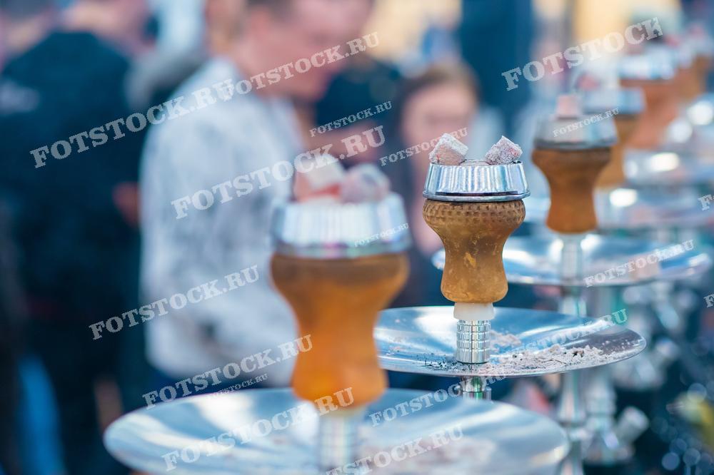 Фотография изображает кальяны с раскаленными углями, готовые к использованию в кальянной. На заднем плане видны люди, наслаждающиеся временем в кальянной, что подчеркивает атмосферу отдыха и общения.